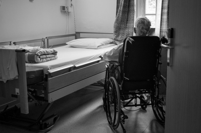 Black-and-white photo of the back of an older man in a wheelchair beside a hospital-type bed.